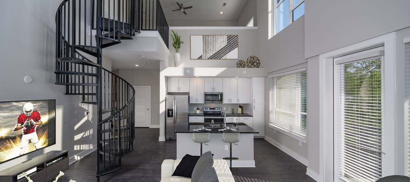 Modern living room with a spiral staircase, a kitchen with a central island, and large windows. A TV plays football, and there is a high ceiling with an upper-level view.