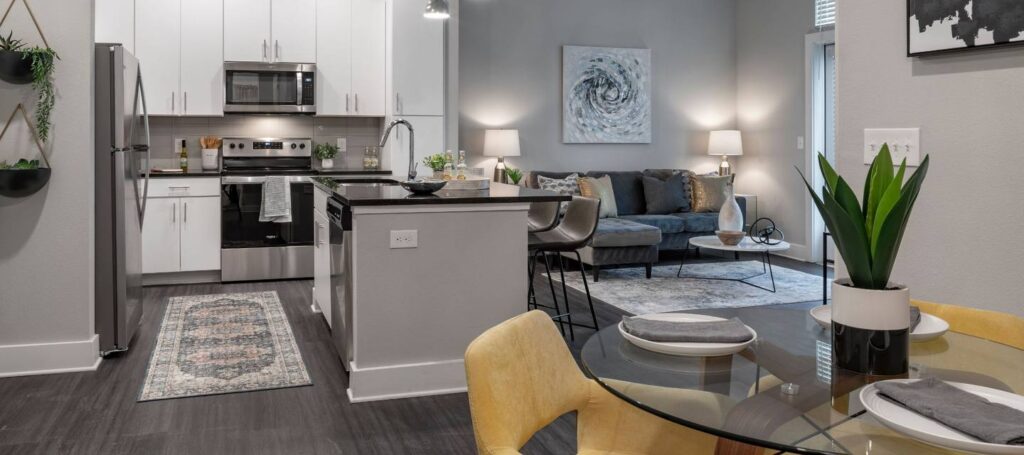 Modern apartment interior with an open kitchen, dining area featuring a round glass table with yellow chairs, and a living room with a gray sofa. Neutral colors and contemporary decor elements.