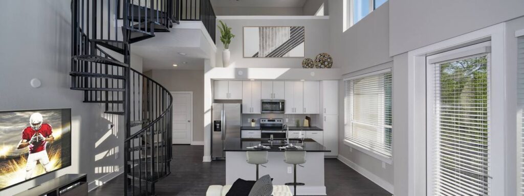 Modern open-concept living space with a spiral staircase, a white kitchen with an island, and large windows. A football game is on the television.