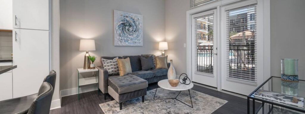 Modern living room with a gray sofa, abstract wall art, two lamps, and a round coffee table. Large windows with blinds offer a view of a balcony and pool area outside.