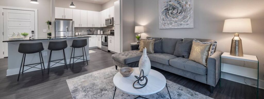 Modern living room and kitchen with gray sofa, white round coffee table, bar stools, and abstract wall art. The space features dark flooring and bright lighting.