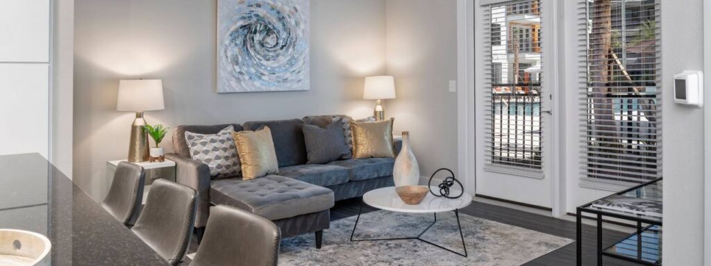 Modern living room with a gray sectional sofa, decorative pillows, a round white coffee table, and a large abstract painting. Two lamps are on side tables, and glass doors lead outside.