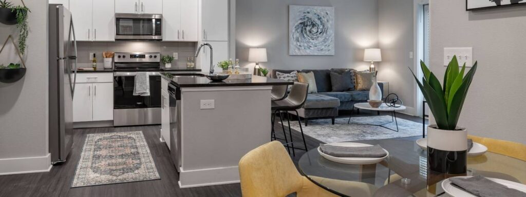 Modern open-concept kitchen and living room with stainless steel appliances, island, gray sofa, abstract art, and decorative plants.