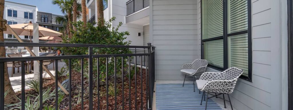 Balcony with two patterned chairs and a striped rug next to a railing. Overlooks a landscaped area with palm trees and patio tables with umbrellas.