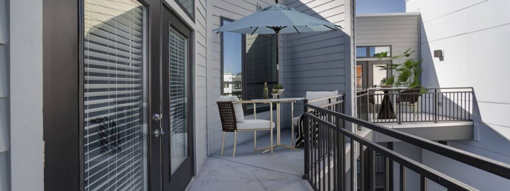 Outdoor balcony with a table, chairs, and umbrella for shade, surrounded by modern railings and building walls, with a small plant in the corner.