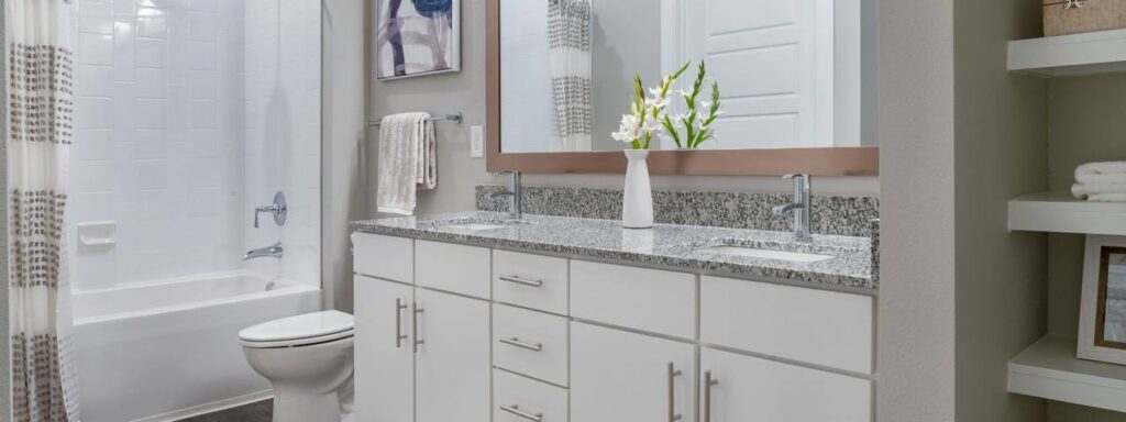 Modern bathroom with a double sink vanity, granite countertop, and white cabinets. A bathtub with a shower curtain is visible, alongside a potted plant and folded towels on display shelves.