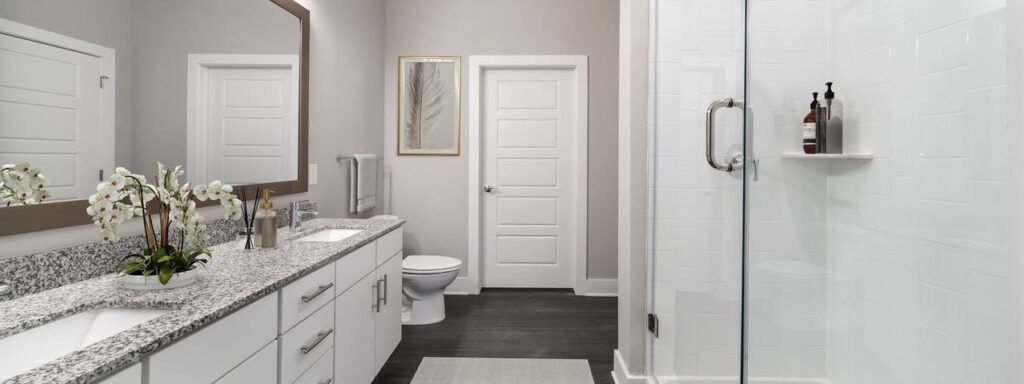 Modern bathroom with a large mirror above a granite countertop, white cabinets, an orchid plant, a glass shower, and a closed door. A framed picture hangs on a light gray wall.