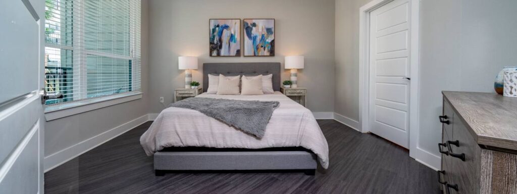 A modern bedroom with a gray bed, white and gray bedding, two abstract paintings, two lamps on side tables, and a chest of drawers. Natural light filters through a large window with blinds.