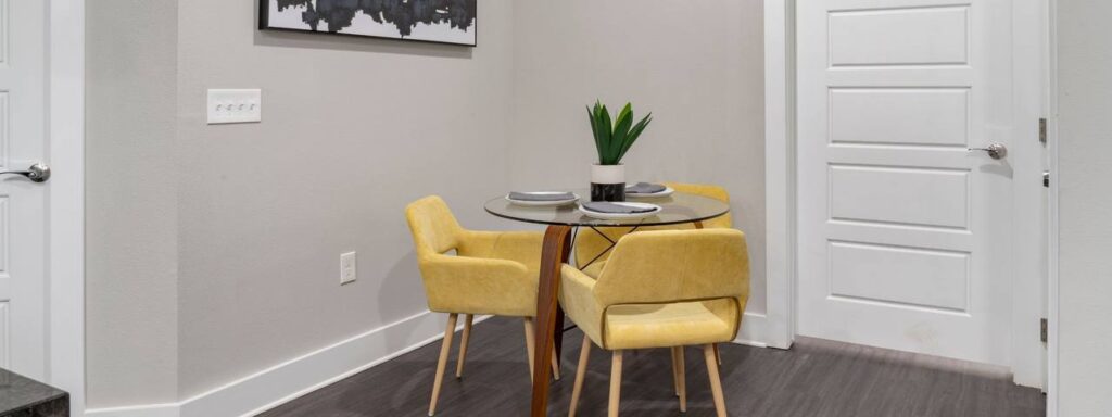 A small dining area with a round glass table, two yellow chairs, a potted plant, and a black and white wall art piece.