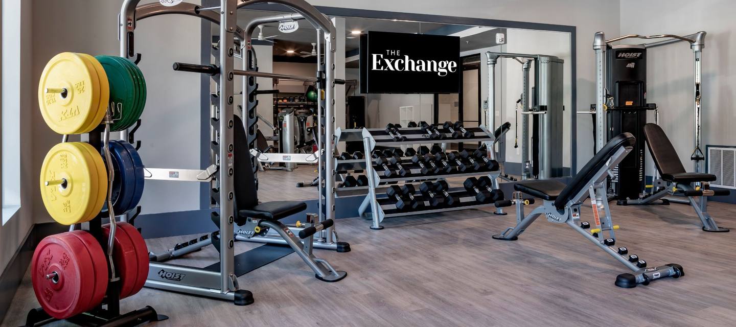 Gym with various weights, benches, and exercise equipment. A mirror reflects the space, and there is a sign reading "The Exchange" on the wall.