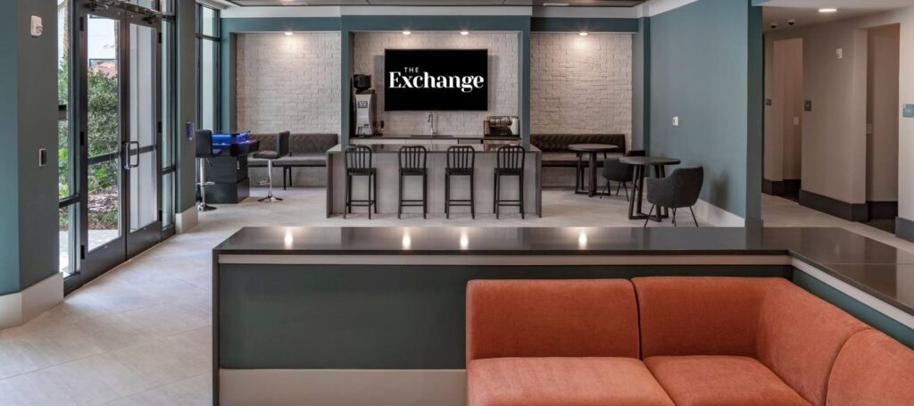 Modern lounge with orange sofa, bar seating, and a screen displaying "The Exchange." Walls are teal and gray with natural light from large windows on the left.