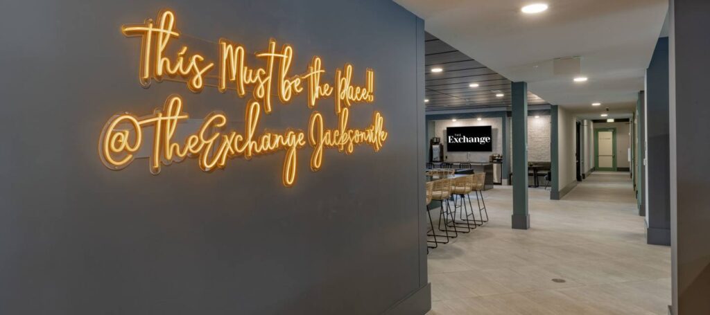 Hallway with neon sign reading "This Must Be The Place! @TheExchangeJacksonville" and seating area with tables, chairs, and a monitor displaying "Exchange.