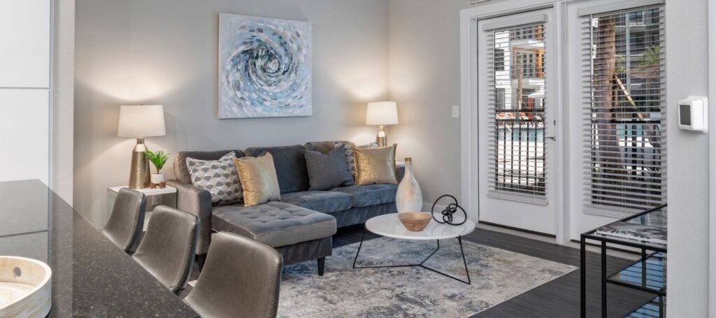 Modern living room with a gray sofa, decorative pillows, abstract wall art, and a round coffee table. Two lamps flank the sofa, and double glass doors with blinds lead outside.