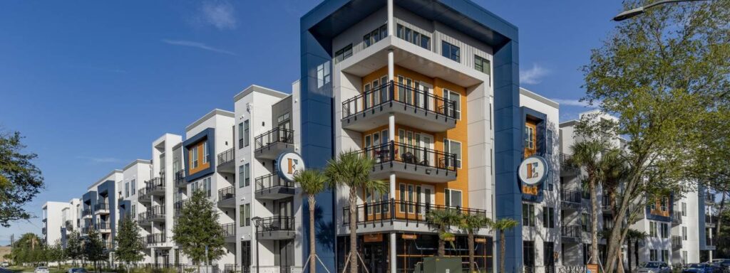 Modern apartment complex with multiple stories, balconies, and a mix of white, blue, and orange accents. Surrounded by trees and a clear blue sky.