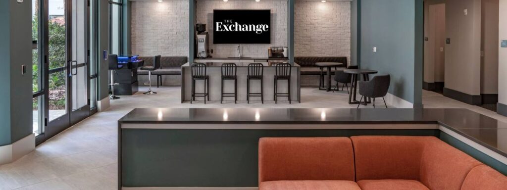 Modern lobby with blue walls, a TV displaying "The Exchange," seating areas, a bar counter with stools, and large windows.