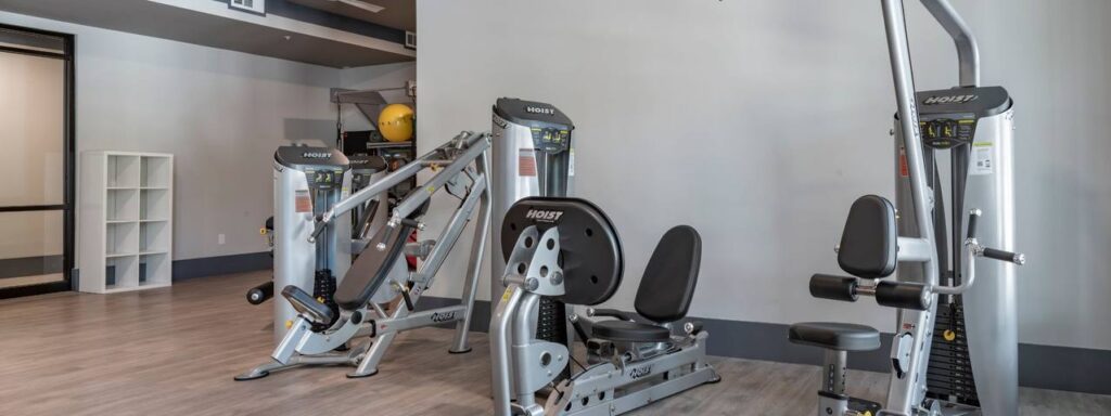 A fitness room with exercise machines, including a seated chest press and a dual adjustable pulley, against a plain wall.