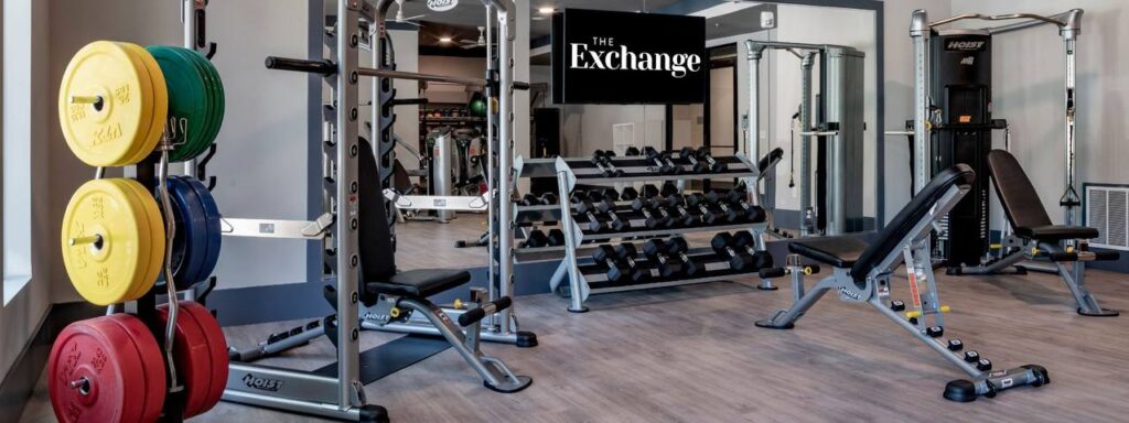 A gym with weight racks, benches, and various workout equipment, featuring a sign that reads "The Exchange.