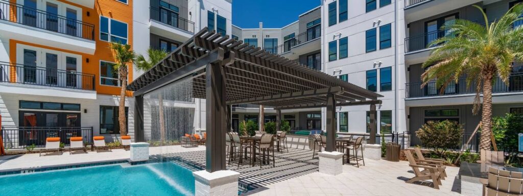 A modern apartment complex features a pool area with lounge chairs, palm trees, and a pergola.