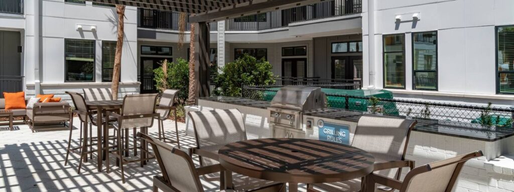 Outdoor patio with tables, chairs, and a grill under a pergola, surrounded by apartment buildings and plants.