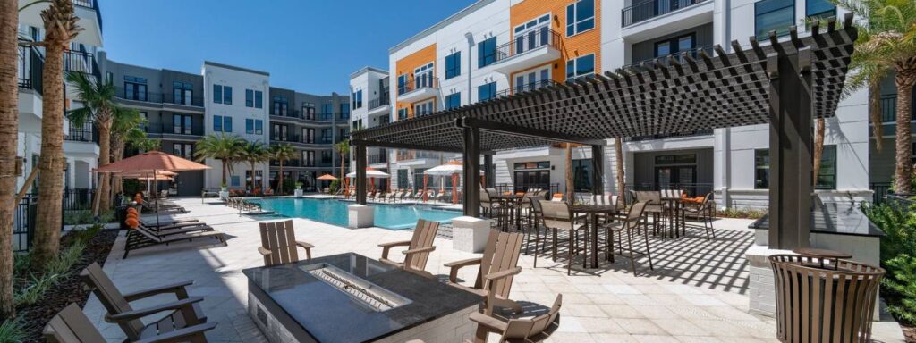 Modern apartment complex with a large outdoor swimming pool, shaded seating areas, lounge chairs, and palm trees.