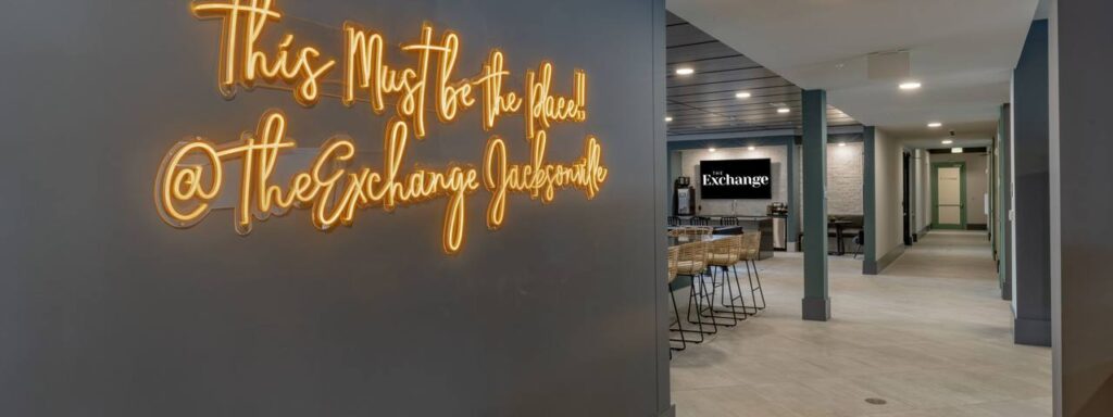 Neon sign reads "This Must be the Place @The Exchange Jacksonville" in a modern room with a long table and chairs.