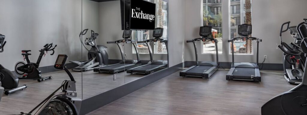 A modern gym with treadmills, stationary bikes, and large mirrored walls. A TV screen displays "The Exchange.