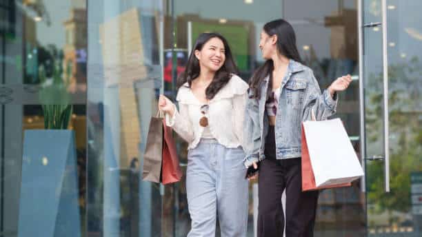 Two women holding shopping bags walk out of a store, smiling and talking to each other.