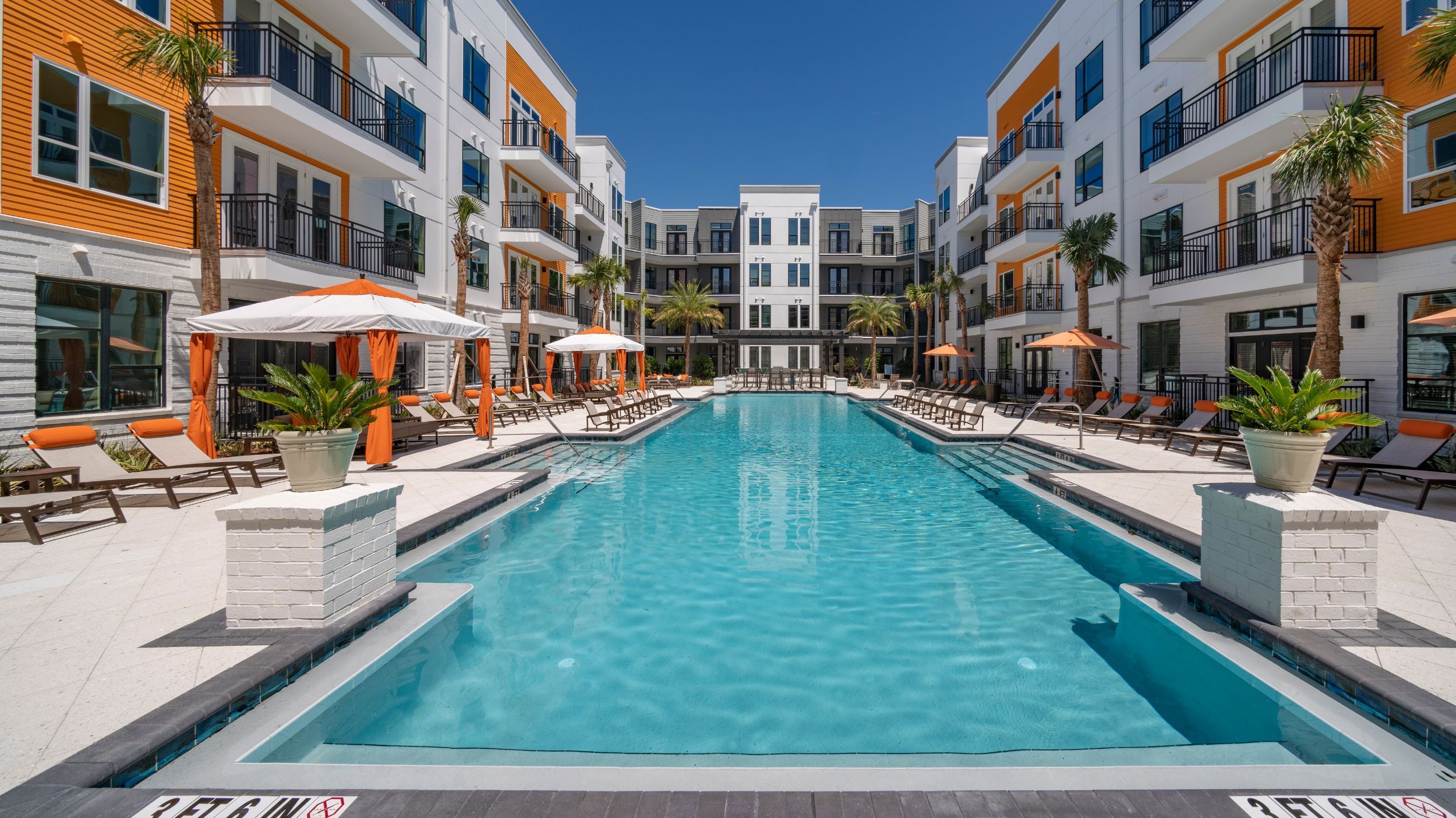 Modern apartment complex with a central outdoor pool surrounded by lounge chairs and umbrellas, framed by three-story buildings under a clear blue sky.