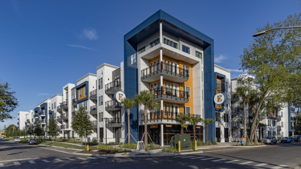 Modern multi-story apartment building with balconies, blue and orange accents, and landscaped surroundings, situated on a corner with clear skies above.