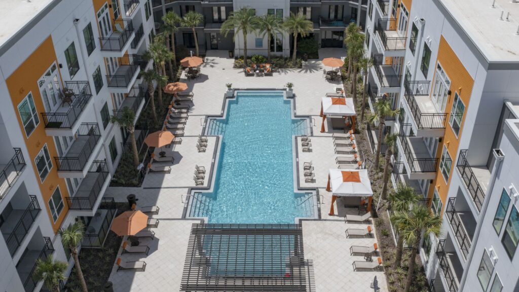 Aerial view of a rectangular outdoor swimming pool surrounded by lounge chairs, umbrellas, and modern apartment buildings. Palm trees are visible around the pool area.