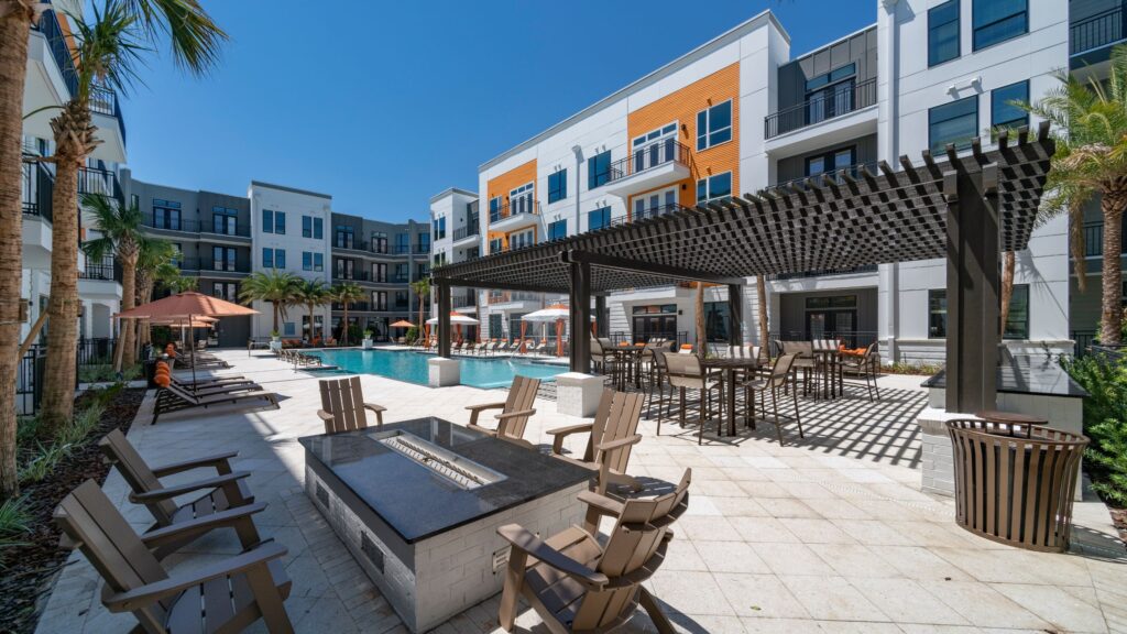 Modern apartment complex with a pool, lounge chairs, fire pit, pergola, and outdoor seating. White and orange building exterior with palm trees. Clear blue sky.