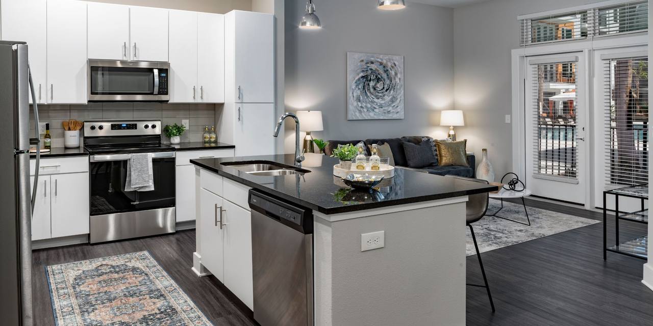 Modern kitchen and living area with a central island, stainless steel appliances, and dark wood flooring. A cozy seating area with a rug and wall art is visible in the background.