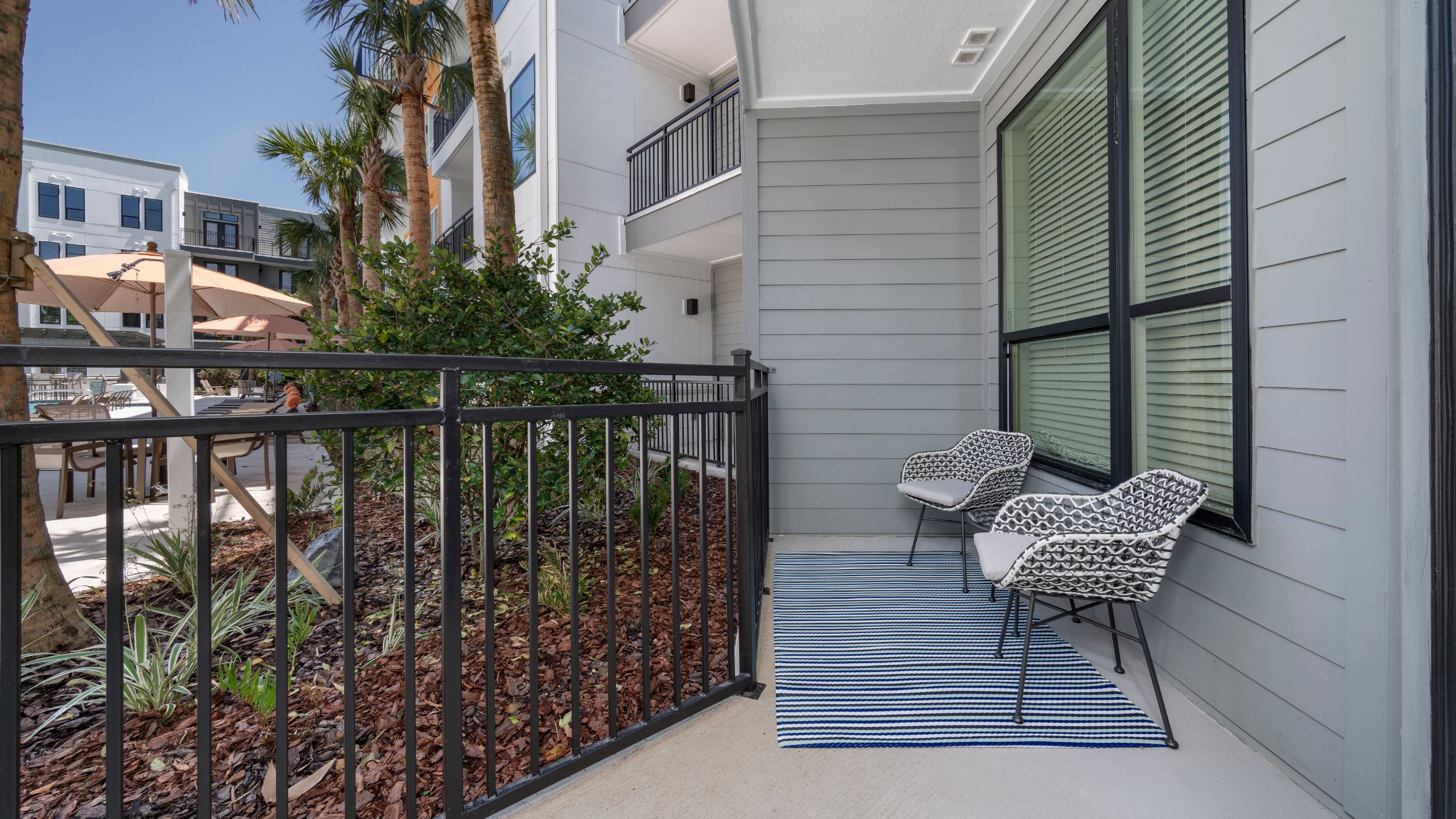 A balcony with two chairs and a striped rug overlooks a landscaped area with a pool and palm trees.