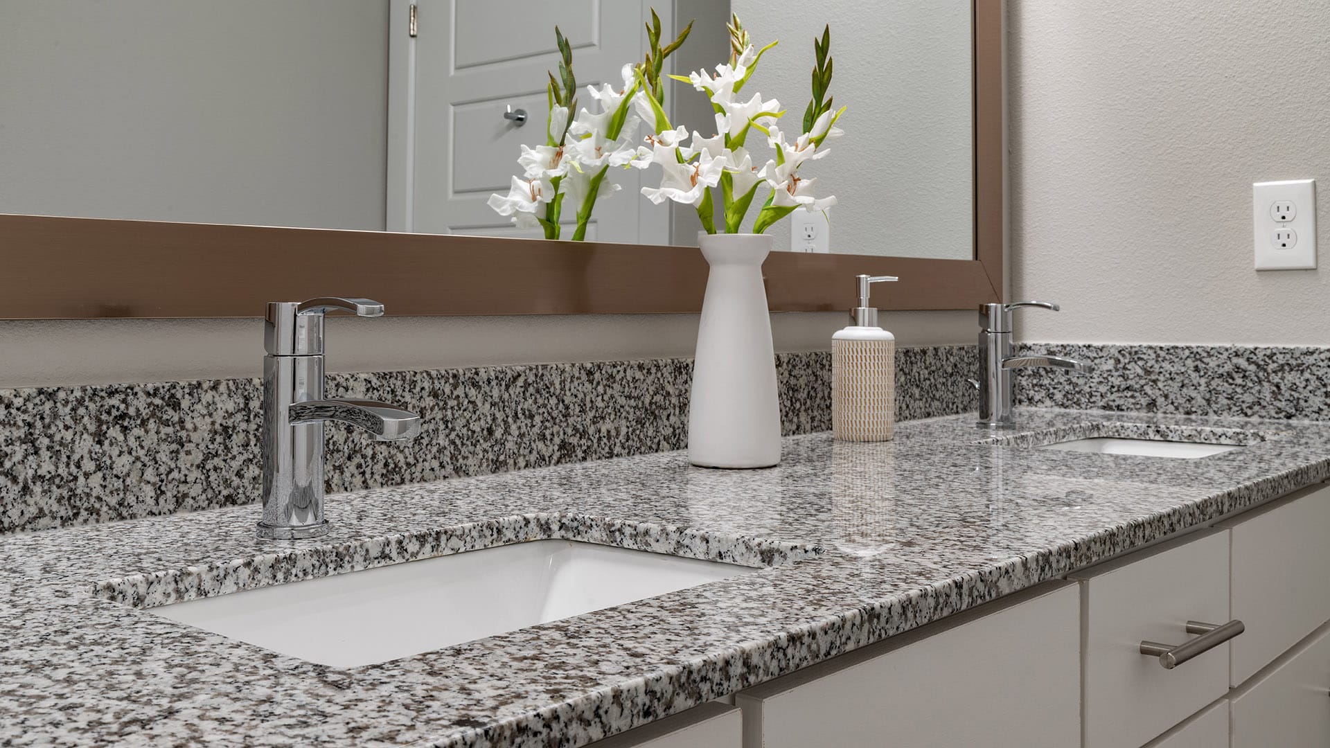 Double-sink bathroom vanity with granite countertop, chrome faucets, a white vase with flowers, a soap dispenser, and a large mirror above.