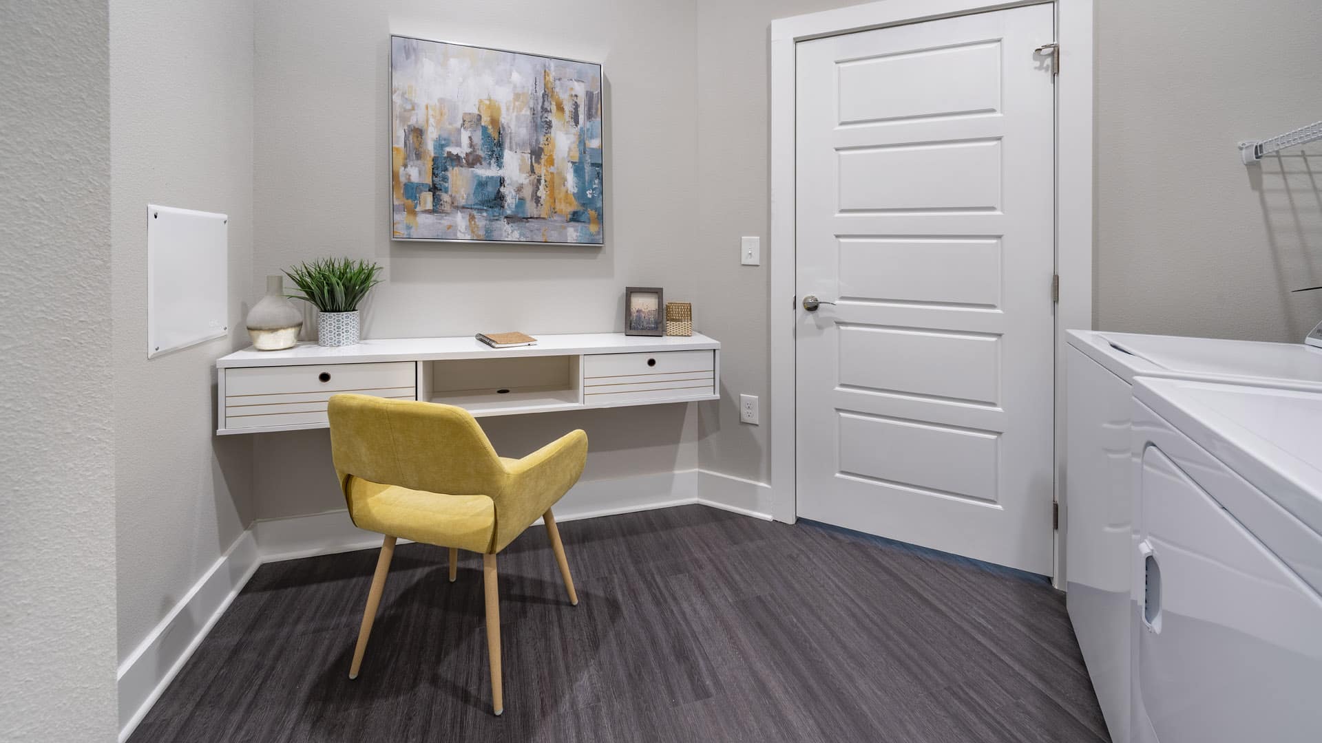 A small home office setup with a yellow chair, white desk, abstract wall art, and a washing machine visible to the right.
